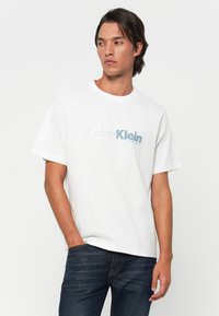 Mand iført hvid Calvin Klein T-shirt og mørke jeans, står med den ene hånd i lommen og kigger sidelæns mod en ensfarvet baggrund.
