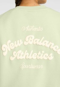 Sweatshirt vert menthe avec lettrage en crème en relief. Le texte comprend "Authentic New Balance Athletics Sportswear" dans une police cursive ludique.