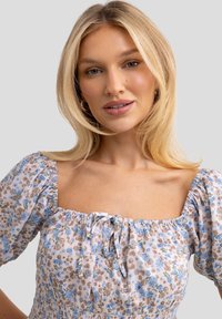 Vestido de tela ligera con estampado floral. Presenta mangas abullonadas y escote cuadrado con cordón ajustable. Los colores incluyen azul, beige y marrón.