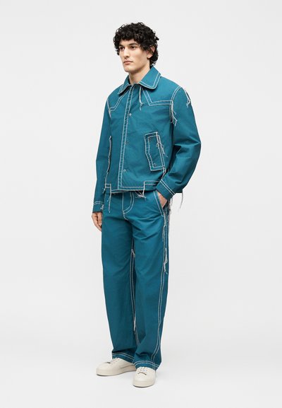 Νέος άνδρας με σγουρά μαλλιά που φοράει τζιν μπουφάν σε πετρόλ χρώμα και ταιριαστό παντελόνι με εμφανείς λευκές ραφές και φθαρμένες άκρες, στέκεται με το ένα χέρι στην τσέπη.