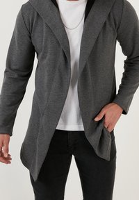 Grå, långärmad kappa med huva, gjord av mjukt tyg, har en draperad framsidan och bärs över en vit skjorta och svarta byxor.