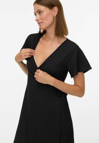 Robe portefeuille noire avec une finition texturée, manches courtes volantées et un décolleté en V profond. Ceinture à nouer à la taille pour un ajustement personnalisé.