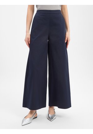 Persoon die marineblauwe wijde broek en zilveren slingback platte schoenen met puntige neus draagt, staand tegen een effen witte achtergrond.