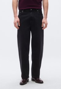 Juodi džinsai iš džinso audinio laisvo kirpimo, su penkiomis kišenėmis, priekyje su sagomis užsegami ir subtiliais siūlių detalėmis. Derinami su tamsiais batais.