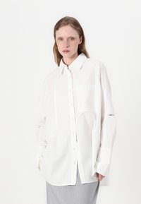 White oversized button-up shirt made of lightweight fabric, featuring a chest pocket and cut-out accents on sleeves.