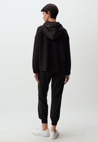 Haut à capuche noir avec un cordon de serrage ajustable, coupe décontractée, poignets côtelés, associé à un pantalon assorti. Des baskets blanches complètent le look.