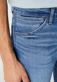 Ljusblå denimjeans med en jämn textur, med orange stygnen, en framficka och en närbild av midjeområdet.