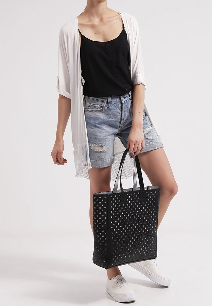 Personne portant un short en jean déchiré bleu clair, un débardeur noir, un cardigan blanc ouvert, des baskets blanches, tenant un grand sac cabas noir avec des pois argentés.
