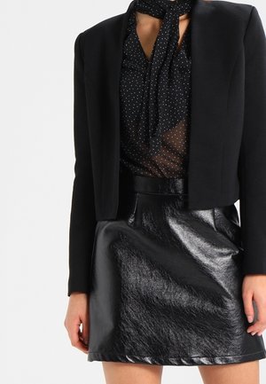 Frau trägt einen schwarzen, kurzen Blazer, eine durchscheinende, gepunktete Bluse mit Schleifenkragen und einen glänzenden schwarzen Leder-Minirock.