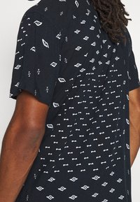 Svart kortärmad skjorta med ett vitt upprepande geometriskt mönster, tillverkad av lättviktig tyg med avslappnad passform.