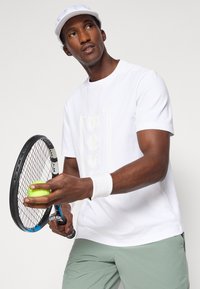 Λευκά κοντομάνικα πουκάμισο με διακριτό ανάγλυφο λογότυπο, κρατώντας μία ρακέτα τένις και μία πράσινη μπάλα, συνδυασμένο με ανοιχτό πράσινο σορτς.