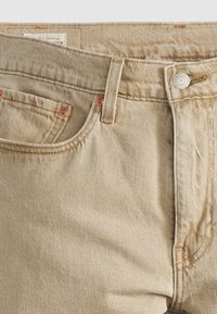 Jean en denim beige en gros plan montrant la poche avant, les passants de ceinture, le bouton en métal, les coutures et la texture du tissu.