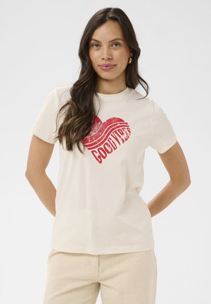 Femme aux longs cheveux bruns portant un t-shirt crème avec un cœur rouge et le texte "GOOD VIBES", assorti à un pantalon beige clair.