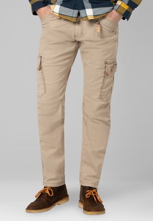 Man draagt beige cargobroek met meerdere zakken, een geruit overhemd ingestopt en bruine leren laarzen met oranje veters. Handen in de zakken.