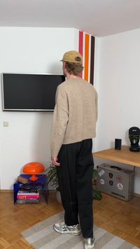 Bež vuneni džemper, tamne hlače, srebrne tenisice. Narančasta lampa na prozirnom plavom noćnom stoliću, TV na zidu, drveni stol s biljkom.