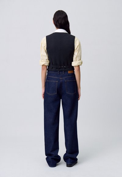 Una persona sta in piedi con la schiena rivolta, indossando un gilet nero sopra una camicia a righe e jeans larghi blu, su uno sfondo grigio chiaro.