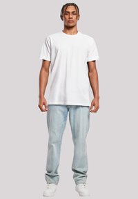 Junger Mann mit geflochtenen Haaren steht frontal, trägt ein schlichtes weißes T-Shirt, hellblaue Jeans und weiße Sneakers.