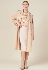Trench coat beige con bottoni doppi, cintura e spalline, sopra un maglione a fantasia e una gonna crema a coste. Stivaletti in suede alla caviglia.