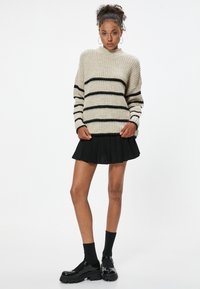Cremfarbener Chunky-Strickpullover mit schwarzen Streifen, ausgestattet mit einem hohen Kragen; kombiniert mit einem schwarzen Faltenrock und schwarzen Plateauschuhen.