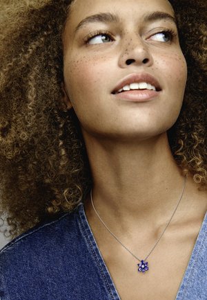 Collana d'argento con un pendente a forma di fiore decorato con pietre blu. La modella indossa un top in denim con scollatura a V, che mette in evidenza la collana.