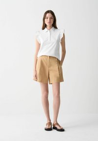 Balta be rankovių polo maikė su volanu ant pečių, derinama su smėlinėmis šortais. Modelis dėvi rudas šlepetes. Šviesus, minimalistinis fonas.