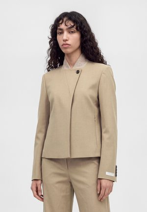 Un mannequin aux longues boucles foncées porte une veste beige cintrée à bouton asymétrique et un pantalon assorti, sur un fond clair uni.