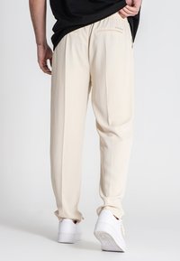 Beige bukser lavet af et blødt materiale, med en afslappet pasform, sidelommer og elastisk talje, parret med hvide sneakers.