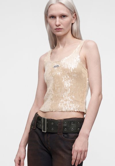 Femme aux longs cheveux blonds portant un haut beige sans manches à sequins et un pantalon foncé avec une large ceinture noire, sur un fond uni clair.