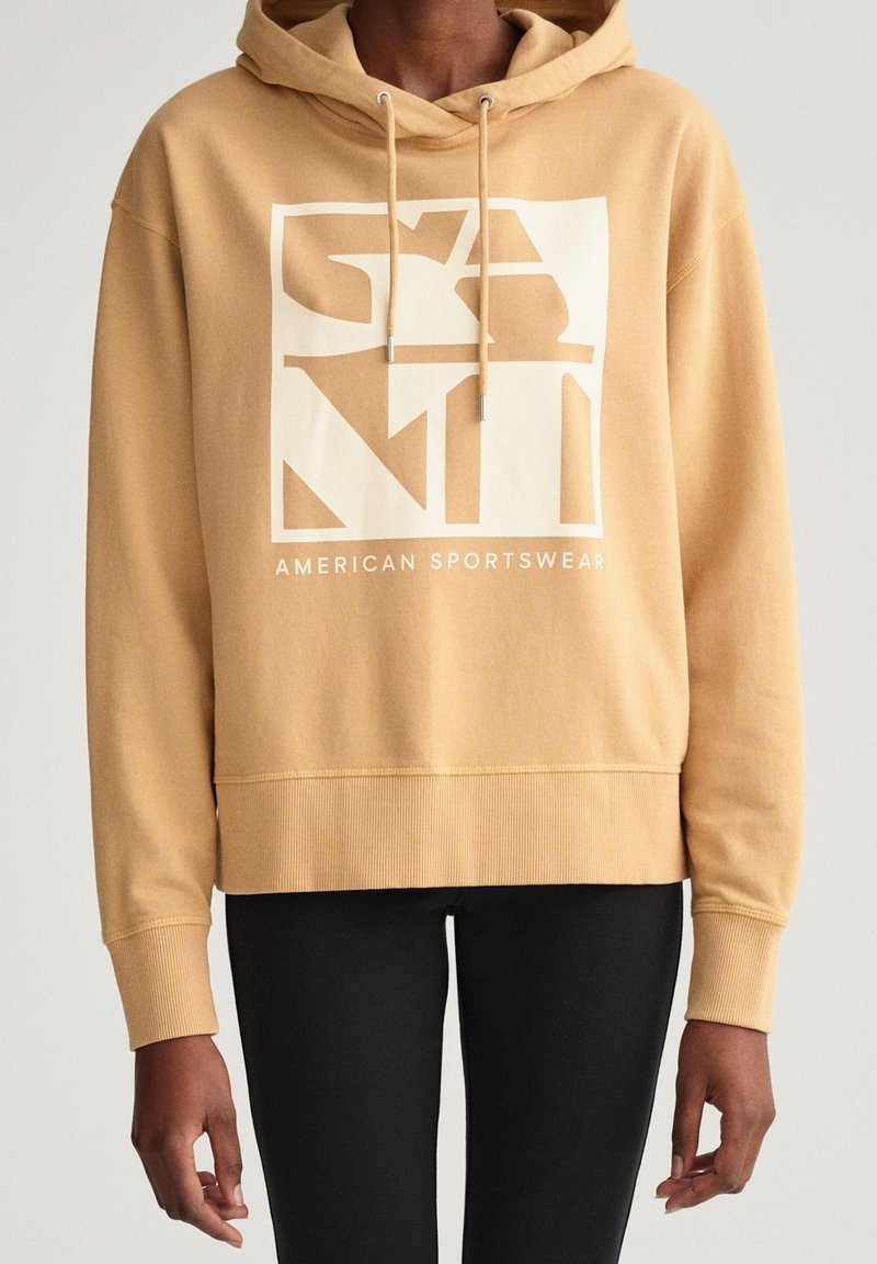 Sweat à capuche beige avec un motif graphique frontal présentant les grandes lettres "GANT" et "AMERICAN SPORTSWEAR", cordons de serrage, poignets et ourlet côtelés.
