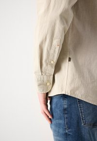 Camicia beige chiaro con polsini abbottonati e fessure laterali, realizzata in un tessuto testurizzato, abbinata a jeans in denim blu.
