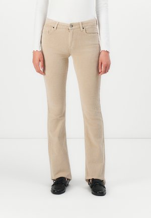 Pantalons en velours côtelé beige portés avec un haut blanc à manches longues et des mocassins à lacets noirs, montrés de la taille aux pieds sur un fond blanc uni.