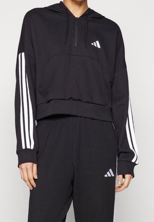 Personne portant un sweat à capuche noir et un pantalon assorti avec des détails blancs à trois bandes sur les manches et les jambes, et un petit logo sur la poitrine et la cuisse.