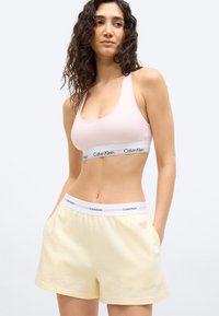 Γυναίκα με σγουρά μαλλιά που φοράει ένα ανοιχτό ροζ αθλητικό σουτιέν Calvin Klein και ανοιχτά κίτρινα σορτς με τα χέρια στις τσέπες, κοιτάζοντας προς τα πλάγια.