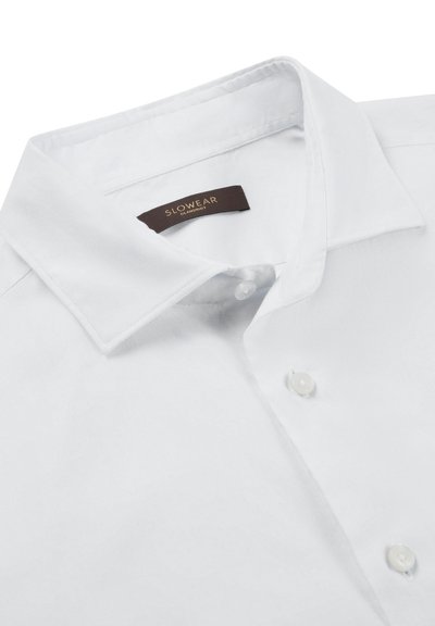 Camisa blanca con cuello, confeccionada en tela lisa con una textura sutil, cierre de botones y una etiqueta marrón de marca en el interior del cuello.