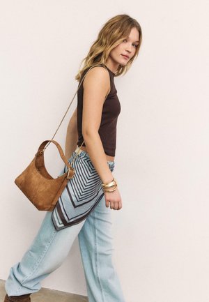 Junge Frau, die einen ärmellosen braunen Oberteil, hellblaue Jeans, goldene Armreifen, einen gemusterten Schal, der an der Taille gebunden ist, und eine braune Leder-Umhängetasche trägt.