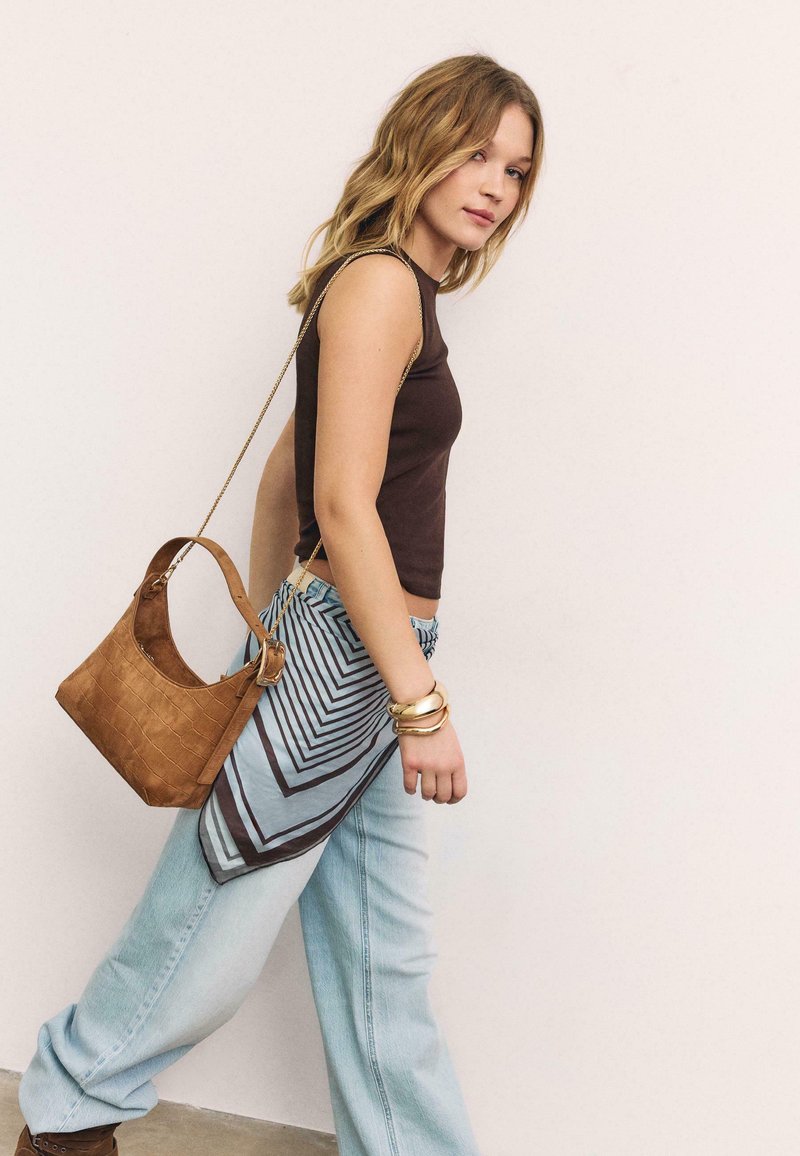 Jeune femme marchant, vêtue d'un haut marron sans manches, d'un jean bleu clair, de bracelets dorés, d'une écharpe à motifs nouée à la taille, et portant un sac à bandoulière en cuir marron.