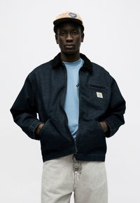 Νεαρός μαύρος άνδρας που φοράει μπεζ καπέλο, σκούρο τζιν μπουφάν με φερμουάρ, ανοιχτό μπλε μπλουζάκι και ανοιχτό γκρι τζιν, στέκεται με τα χέρια στις τσέπες.