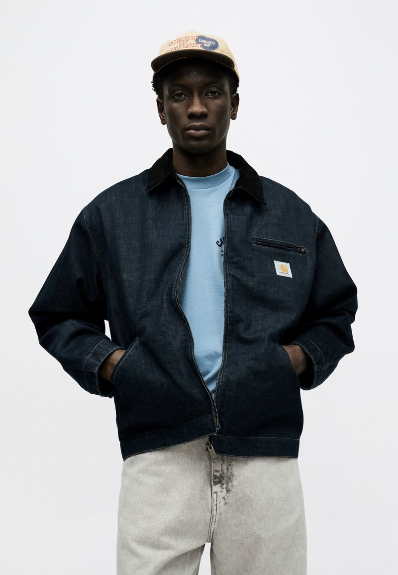 Joven hombre negro con gorra beige, chaqueta de mezclilla oscura con cremallera, camiseta azul claro y vaqueros gris claro, de pie con las manos en los bolsillos.