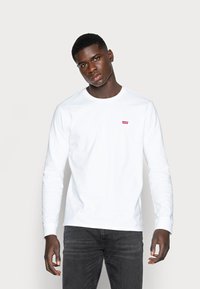 White long-sleeve t-shirt made of cotton with a small red Levi's logo on the chest. Worn with dark gray jeans.