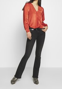 Blouse en satin rouge avec col en V et manches longues, associée à un jean noir évasé et une ceinture avec des accents métalliques. Bottes à motif peau de serpent.