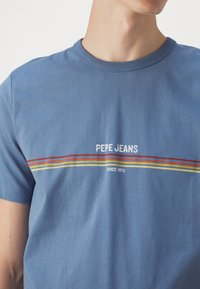 Kék pamut póló, amely a "PEPE JEANS" logót és vízszintes csíkokat tartalmaz piros, narancssárga és sárga színekben, a logo alatt pedig a "SINCE 1973" felirat található.