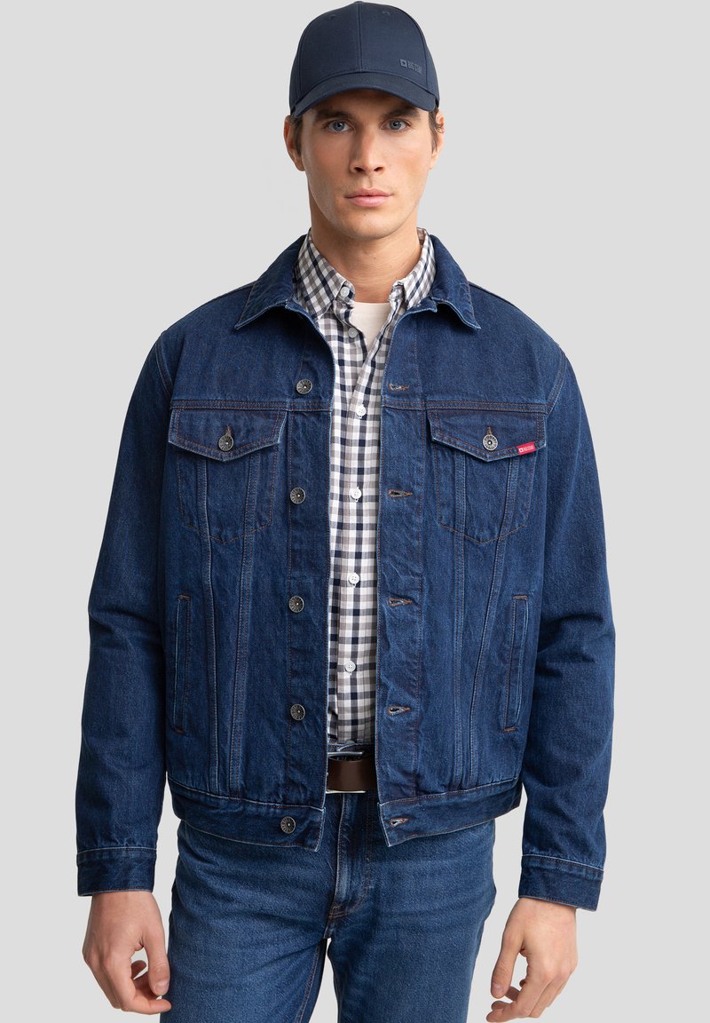 Modra denim jakna z dvema žepoma na prsih, gumbnimi zapiranji in klasičnim ovratnikom, prekrita z karo srajco in temnimi kavbojkami.