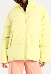 Chaqueta acolchada amarilla con cierre de cremallera, acolchado cosido horizontalmente y bolsillos laterales. Textura suave, corte oversize y cuello alto.