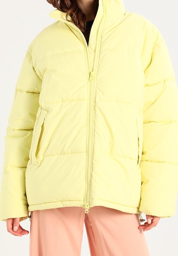 Chaqueta acolchada amarilla con cierre de cremallera, acolchado cosido horizontalmente y bolsillos laterales. Textura suave, corte oversize y cuello alto.