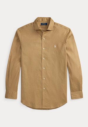 Cămașă bej cu mâneci lungi, cu nasturi, guler, logo alb brodat pe piept și manșete cu nasturi, așezată plat pe un fundal alb.