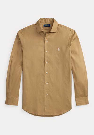 Μπεζ πουκάμισο με μακριά μανίκια, κουμπιά και γιακά, λευκό κεντημένο λογότυπο στο στήθος και μανσέτες με κουμπιά, απλωμένο επίπεδα σε λευκό φόντο.