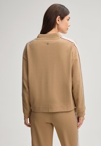 Felpa marrone a maniche lunghe con colletto alto e strisce laterali a contrasto. Tessuto liscio con un piccolo logo sulla parte posteriore del collo.