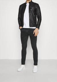 Man som bär en svart läderjacka, vit T-shirt, mörkgrå jeans och vita sneakers står mot en enkel ljus bakgrund.