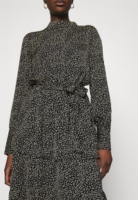 Femme portant une robe noire à col montant et manches longues avec un motif de points irréguliers blancs, ceinture nouée à la taille, et boucles d'oreilles créoles dorées.