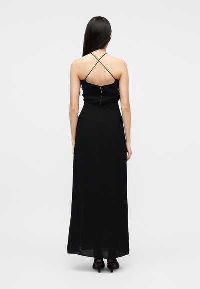 Γυναίκα με μακριά σκούρα μαλλιά, φοράει μαύρο αμάνικο μάξι φόρεμα με λεπτές λουρίδες που διασταυρώνονται στην πλάτη και μαύρα ψηλοτάκουνα, στέκεται μπροστά σε λευκό φόντο.